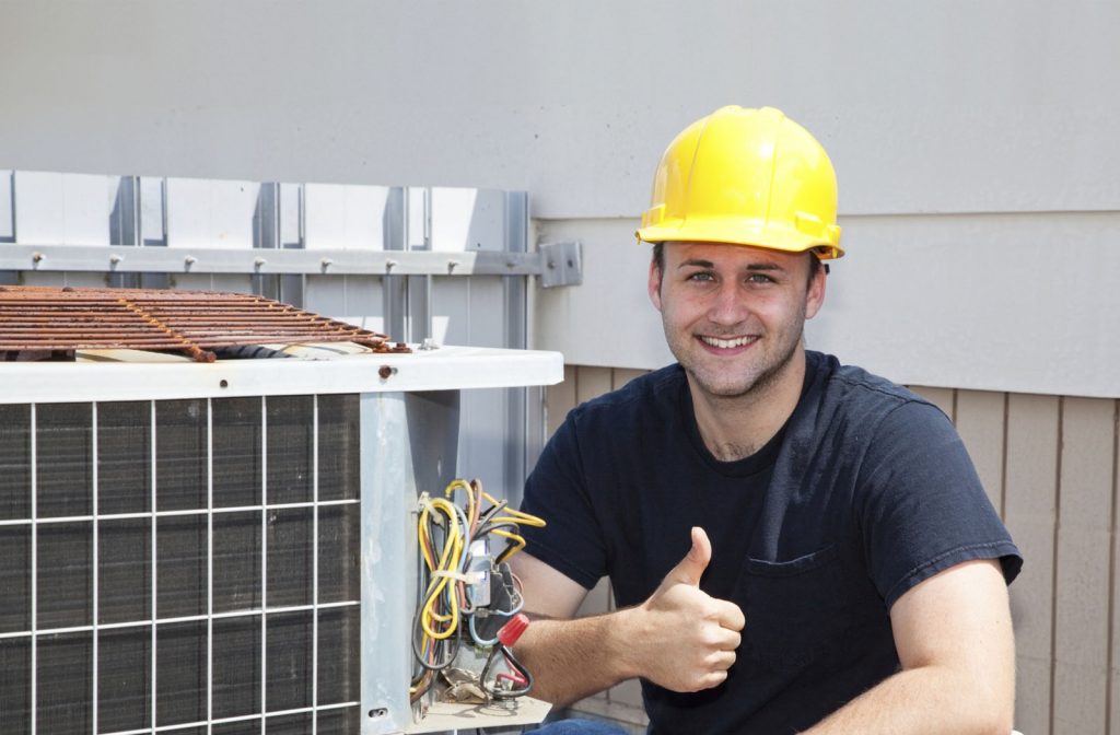 HVAC contractor posting next to an AC unit, smiling and giving a thumbs-up